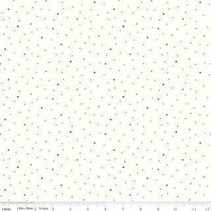 Op de afbeelding: Een witte stof met een verspreid patroon van kleine zwarte stippen van verschillende groottes. Aan de onderkant is een liniaal zichtbaar met metingen in centimeters van 0 tot 30. De tekst Riley Blake Designs staat onderaan gedrukt.