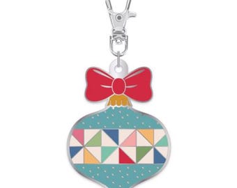 May include: A decorative Christmas ornament-shaped charm with a silver clasp. The ornament is teal with a geometric pattern of colorful triangles and a red bow at the top. The charm is designed for use as a keychain or pendant.