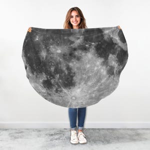 May include: A round, black and white throw blanket with a detailed image of the moon's surface. The blanket is being held up by a person.