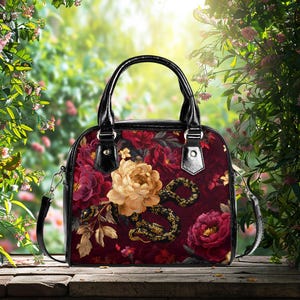 Peut inclure: Sac à main carré avec un motif floral bordeaux, crème et or. Le sac est doté d'une poignée et d'une bandoulière noires. Un motif de serpent doré et noir orne le devant du sac.