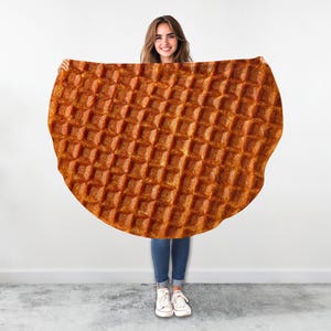 May include: A large, round blanket with a waffle design. The blanket is a warm, golden brown color and features a repeating square pattern. The blanket is held up by a person in blue jeans and white sneakers.