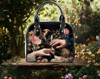 Può includere: Borsa nera con manico superiore e illustrazione dettagliata. L'opera d'arte presenta mani, funghi e fiori in un ambiente da giardino. La borsa ha una finitura lucida e una forma strutturata.