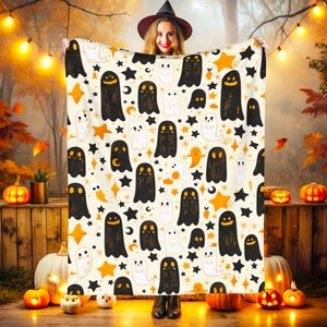 Op de afbeelding: Een crèmekleurige Halloween-deken met een herhalend patroon van zwarte en witte geesten, oranje sterren en zwarte halve manen. De deken wordt omhoog gehouden, waarbij het ontwerp wordt getoond. De achtergrond omvat pompoenen en herfstbladeren.