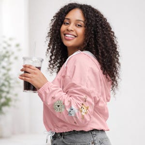 Könnte beinhalten: Hellrosa Kapuzenjacke mit gestickten Katzenmotiven auf dem linken Ärmel. Die Jacke hat einen weißen Kordelzug und ein weißes Futter in der Kapuze. Die Person hält einen transparenten Plastikbecher mit einem Strohhalm.