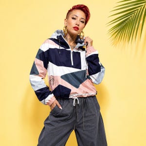 Könnte beinhalten: Eine langärmelige Windjacke zum Überziehen mit einem geometrischen Color-Block-Muster in Marineblau, Weiß, Hellrosa und Anthrazit. Die Jacke verfügt über einen halben Reißverschluss, eine Kapuze mit Kordelzug und eine kleine Gesichtsgrafik.