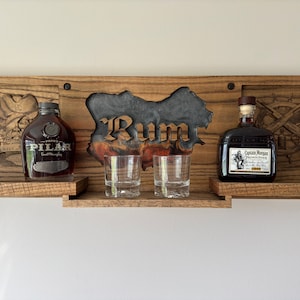 May include: Wooden wall shelf with two bottles of rum, Captain Morgan and  Pilar, and two glasses. The shelf has a carved design of a skull and crossbones, a ship's wheel, and the word "Rum".