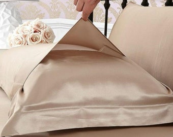 Puede incluir: Primer plano de una funda de almohada de satén beige que se levanta de una cama. La funda de almohada es lisa y sedosa, con un brillo sutil.