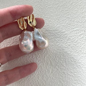 Puede incluir: Un par de pendientes dorados con grandes perlas irregulares de color blanquecino. Los pendientes tienen una parte superior dorada texturizada. Las perlas tienen un brillo lustroso e iridiscente. Los pendientes son sostenidos por una mano sobre un fondo blanco texturizado.