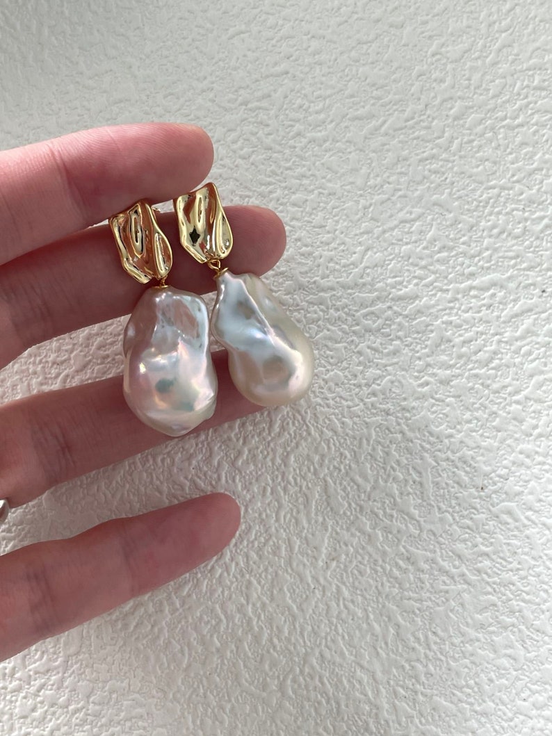 Puede incluir: Un par de pendientes dorados con grandes perlas irregulares de color blanquecino. Los pendientes tienen una parte superior dorada texturizada. Las perlas tienen un brillo lustroso e iridiscente. Los pendientes son sostenidos por una mano sobre un fondo blanco texturizado.