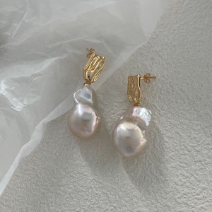 Puede incluir: Un par de pendientes dorados con grandes perlas blancas de forma irregular. Los pendientes tienen una secci&oacute;n superior dorada texturizada. El fondo es de un color claro y neutro, resaltando el dise&ntilde;o de la joyer&iacute;a.