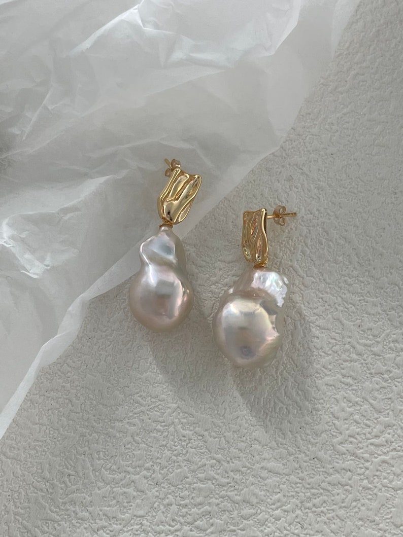Puede incluir: Un par de pendientes dorados con grandes perlas blancas de forma irregular. Los pendientes tienen una secci&oacute;n superior dorada texturizada. El fondo es de un color claro y neutro, resaltando el dise&ntilde;o de la joyer&iacute;a.