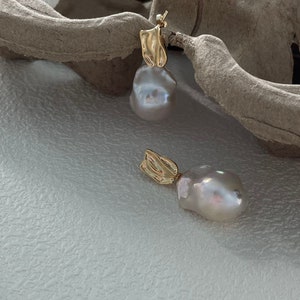Puede incluir: Un par de pendientes dorados con un dise&ntilde;o &uacute;nico. Cada pendiente presenta una perla grande, de forma irregular, de color blanquecino, suspendida de un elemento dorado texturizado. Los pendientes se muestran sobre una superficie blanca texturizada.