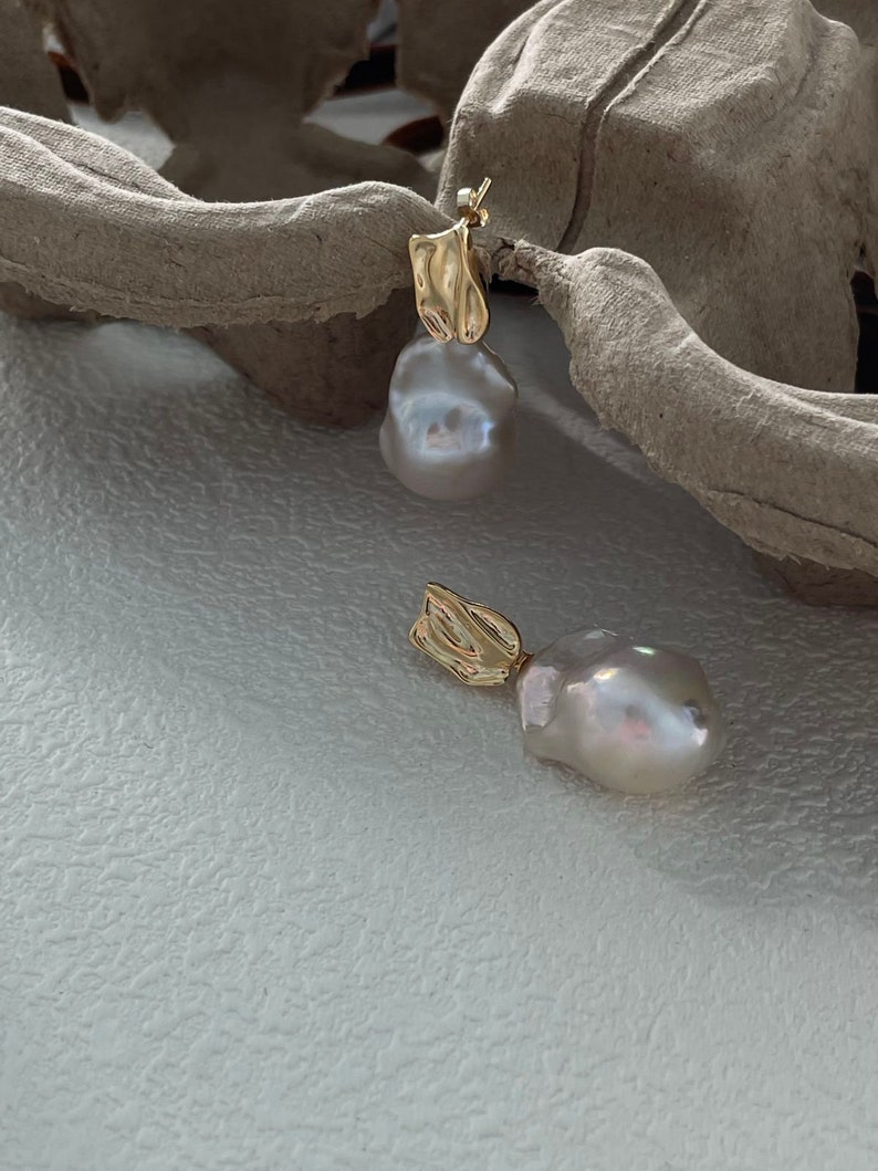 Puede incluir: Un par de pendientes dorados con un dise&ntilde;o &uacute;nico. Cada pendiente presenta una perla grande, de forma irregular, de color blanquecino, suspendida de un elemento dorado texturizado. Los pendientes se muestran sobre una superficie blanca texturizada.