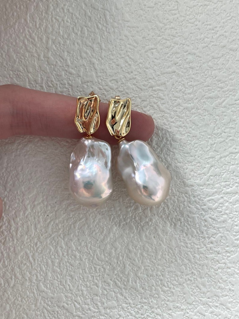 Puede incluir: Un par de pendientes dorados con grandes perlas blancas de forma irregular. Los pendientes tienen una parte superior dorada texturizada con un dise&ntilde;o &uacute;nico. Las perlas tienen un brillo iridiscente y brillante.