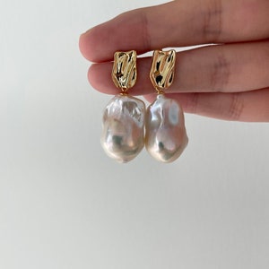 Puede incluir: Un par de pendientes dorados con una parte superior rectangular texturizada y una gran gota de perla blanca irregular. Los pendientes se muestran sobre un fondo blanco liso.