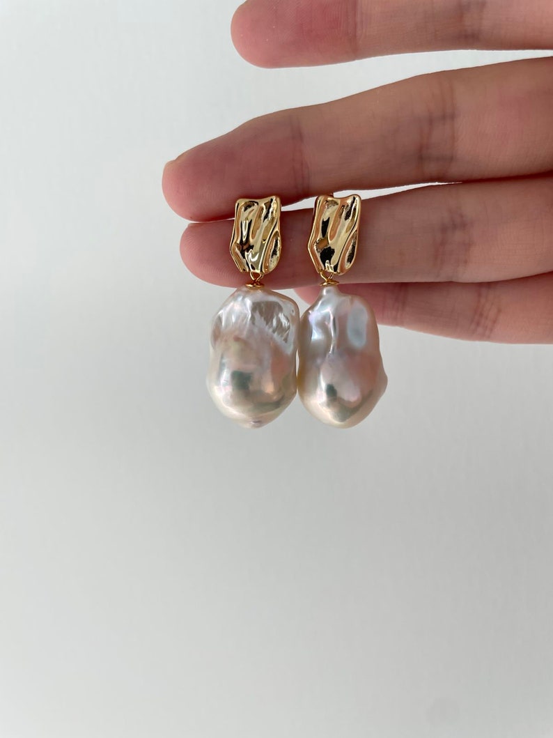 Puede incluir: Un par de pendientes dorados con una parte superior rectangular texturizada y una gran gota de perla blanca irregular. Los pendientes se muestran sobre un fondo blanco liso.