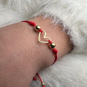 May include: Red string bracelet with a gold heart charm and gold beads.