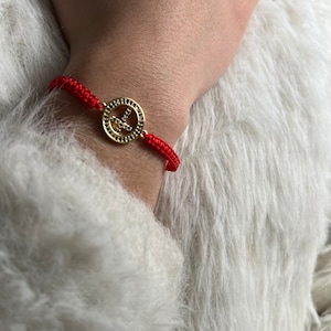 May include: Red string bracelet with a gold peace sign charm. The charm is encrusted with small clear stones.