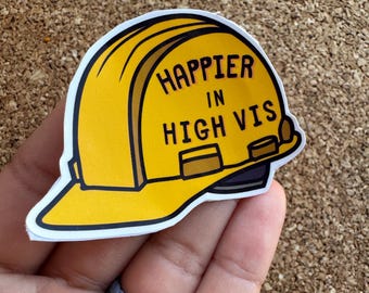 Puede incluir: Una pegatina amarilla de casco de trabajador de la construcción con el texto "Happier in High Vis".