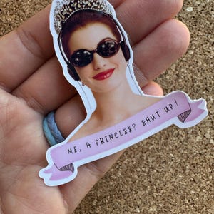 May include: A sticker featuring a woman wearing a tiara and sunglasses, with headphones on. The sticker has a pink banner with the text "ME, A PRINCESS?? SHUT UP!"