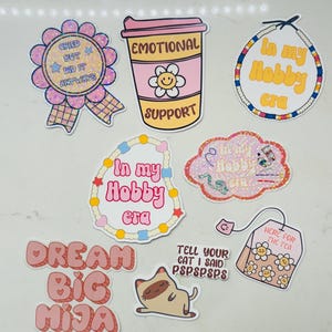 May include: A set of nine stickers with various designs and phrases. The stickers are pink, white, and yellow with glitter accents. The phrases on the stickers include "Cried But Did It Anyways", "Emotional Support", "In My Hobby Era", "Dream Big Mija", "Tell Your Cat I Said PSPSPSPS", and "Here For The Tea".