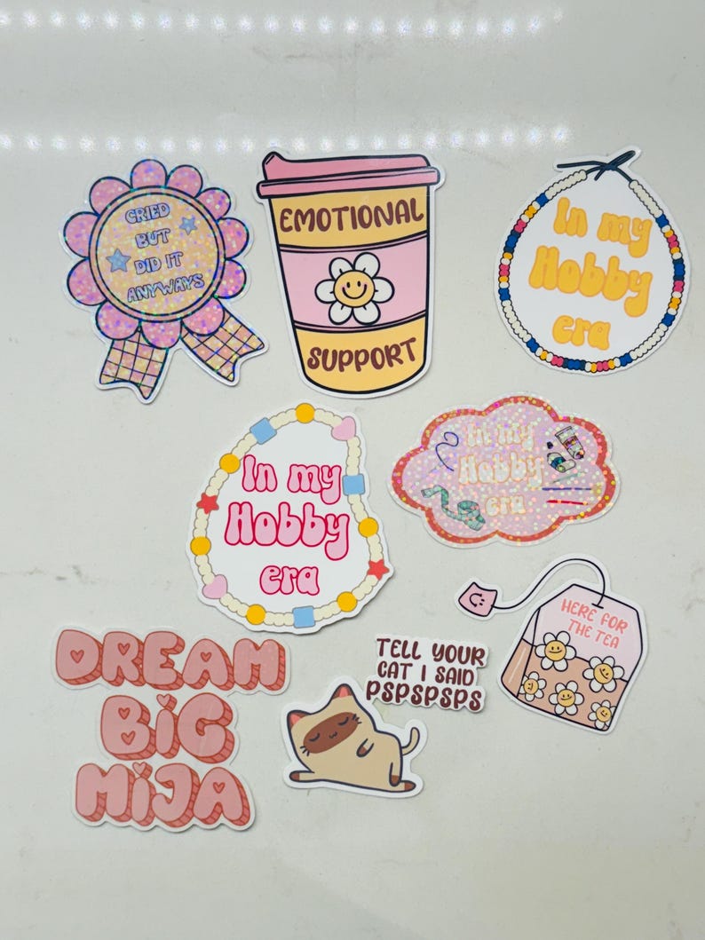 May include: A set of nine stickers with various designs and phrases. The stickers are pink, white, and yellow with glitter accents. The phrases on the stickers include "Cried But Did It Anyways", "Emotional Support", "In My Hobby Era", "Dream Big Mija", "Tell Your Cat I Said PSPSPSPS", and "Here For The Tea".