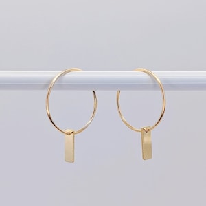 Könnte beinhalten: Ein Paar goldene Creolen mit einem kleinen, rechteckigen Anhänger an jeder Creole.