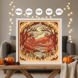 Peut inclure: Une œuvre d'art en papier en couches encadrée représentant un paysage d'automne avec des arbres, une rivière et des montagnes. L'œuvre utilise des tons orange, marron et jaune. Le cadre est blanc et l'image contient le texte "PNG", "SVG", "PDF", "DXF" et "EPS".