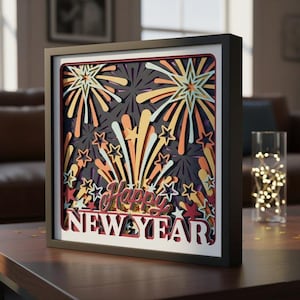 May include: A framed, layered paper art piece with a black frame. The artwork features a fireworks display in shades of orange, yellow, and red against a dark gray background. The words "Happy New Year" are prominently displayed.