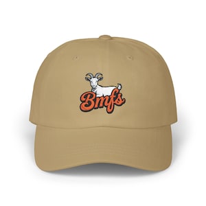May include: A beige baseball cap with a curved peak. The cap has an embroidered design of a goat and the word "Bmfs" in orange and white script. The hat is made of a soft material.