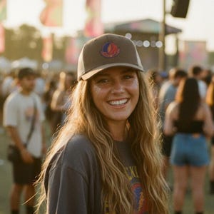 May include: A woman smiles, wearing a brown cap with a logo, a grey t-shirt, and denim shorts. The image is taken outdoors at a festival, with a crowd and stage in the background. The cap has a colorful logo.
