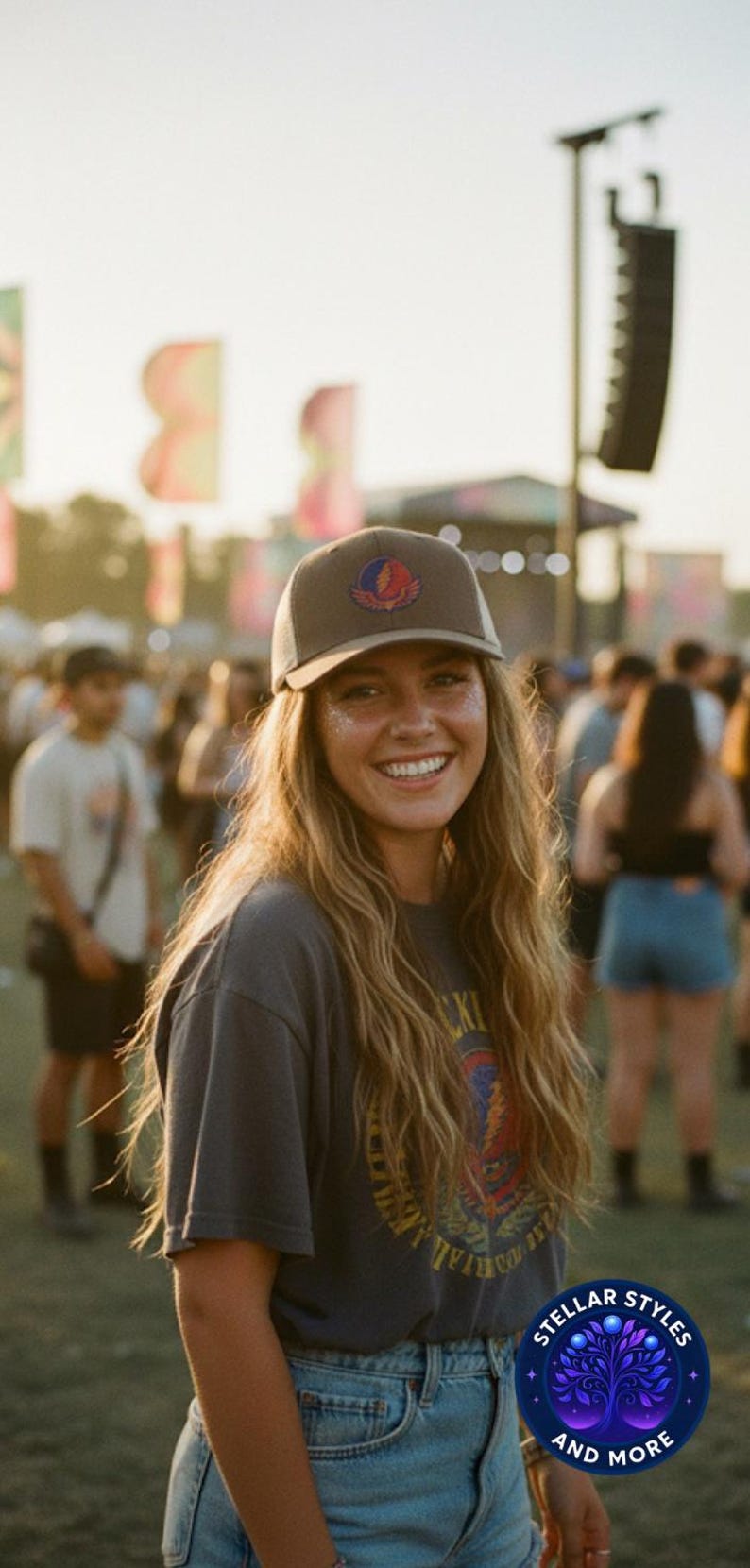 May include: A woman smiles, wearing a brown cap with a logo, a grey t-shirt, and denim shorts. The image is taken outdoors at a festival, with a crowd and stage in the background. The cap has a colorful logo.