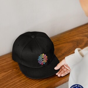 Peut inclure: Casquette de baseball noire à visière plate. La casquette présente un motif floral coloré avec les lettres "LSD" brodées au centre. Le motif incorpore un arc-en-ciel de couleurs. La casquette est posée sur une surface en bois.