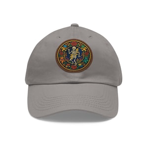 Könnte beinhalten: Graue Baseballkappe mit einem kreisförmigen Aufnäher mit einem tanzenden Skelett, einem Blitz und floralen und Bärenmotiven. Der Aufnäher hat einen braunen Rand. Die Kappe hat einen gebogenen Schirm und einen Stoffriemen.
