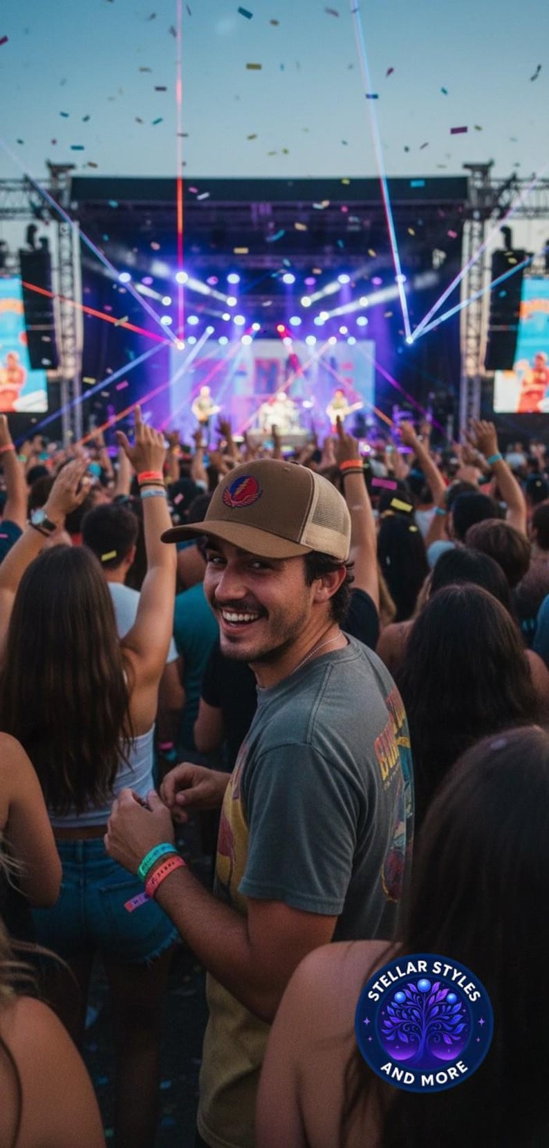 May include: A man smiles at the camera at a concert, wearing a brown trucker hat with a logo and a grey t-shirt. The background shows a stage with lights, a crowd, and confetti.