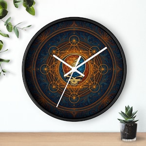 May include: Circular black wall clock with a blue and orange geometric pattern, featuring a skull and lightning bolt emblem. The clock has white hands and is displayed on a white wall. The background includes a plant.