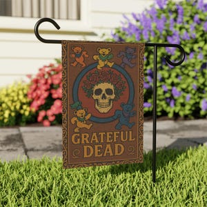 Puede incluir: Una bandera de jardín marrón con una calavera con una corona floral y osos bailarines. La bandera tiene las palabras "Grateful Dead". La bandera se muestra en un soporte de metal negro en un jardín con césped.