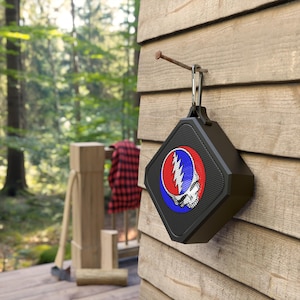 May include: A black portable Bluetooth speaker with a Grateful Dead logo on the front. The speaker is hanging from a nail on a wooden wall.