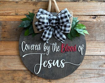 May include: A round wooden sign with the text "Covered by the Blood of Jesus" in white and red lettering. The sign is decorated with a black and white checkered bow, burlap, and green leaves.