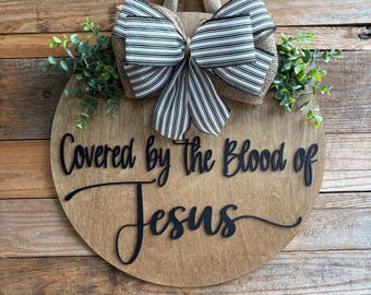 May include: A round wooden sign with the text "Covered by the Blood of Jesus" in black script. It features a burlap bow with black and white striped ribbon and greenery. The sign hangs from a burlap loop.