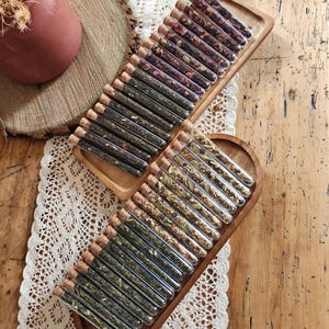 Op de afbeelding: Twee houten trays gevuld met glazen reageerbuizen met verschillende gedroogde kruiden en bloemen. De reageerbuizen zijn voorzien van kurken stoppen. De trays liggen op een wit kantkleed.