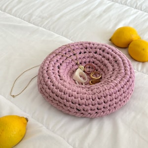 Può includere: Una ciotola rosa all'uncinetto con una collana a catena dorata e anelli al suo interno. La ciotola è su un copriletto bianco con due limoni accanto.