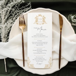 May include: A white menu card with gold accents and a gold crest with the initials "EK" on a white plate with gold cutlery. The menu includes starters, main courses, dessert, and drinks. The text on the menu reads "The Menu", "Appetizers", "Garden Fresh Caprese Skewers", "Mini Chicken Cordon Bleu Bites Avocado", "Entrees", "Atlantic Salmon Seared and Topped with White Wine, Diced Pineapple, Sweet Red Pepper and Onion", "Dessert", "Chocolate Cake with Custard Filling", "Creme Brulee", "Custard Pudding with Fresh Fruit", "Beverages", "Classic Champagne Toast", "Mint and Cucumber Infused Water", "Berry Bliss Sangria", and "Enjoy!"