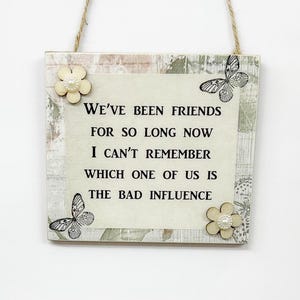 Könnte beinhalten: Ein weißes Holzschild mit einer abgenutzten Oberfläche zeigt einen schwarz-weißen Schmetterling und den Text "WE'VE BEEN FRIENDS FOR SO LONG NOW I CAN'T REMEMBER WHICH ONE OF US IS THE BAD INFLUENCE".