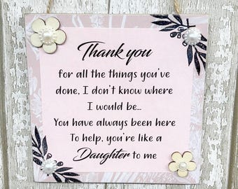 May include: A pink and white wooden sign with black text that reads "Thank you for all the things you've done, I don't know where I would be... You have always been here To help, you're like a Daughter to me". The sign has white flowers with pearl centers and black floral accents.