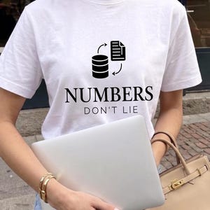 May include: A white short-sleeved t-shirt featuring a black graphic of a database icon and a document icon with arrows, accompanied by the black text "NUMBERS DON'T LIE" printed in a bold serif font across the center of the chest area.