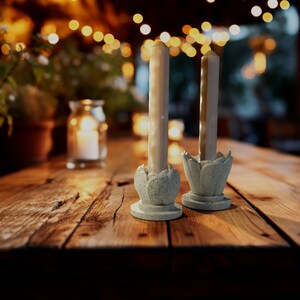 May include: Two white candles in simple, light blue, flower-shaped candle holders on a wooden table. The candles are lit and the table is in a dimly lit setting.
