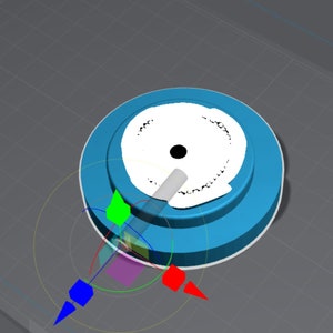 May include: A blue and white circular object with a white center and a black dot in the center. The object is on a gray surface with a grid pattern.