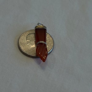 May include: A red carnelian gemstone pendant with a silver setting. The pendant is shaped like a hand with a pointed finger. The pendant is about the size of a US quarter.