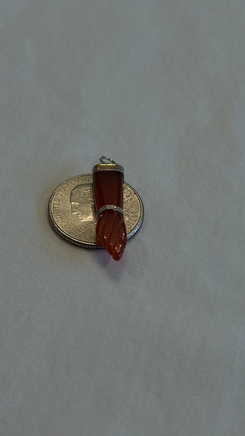 May include: A red carnelian gemstone pendant with a silver setting. The pendant is shaped like a hand with a pointed finger. The pendant is about the size of a US quarter.